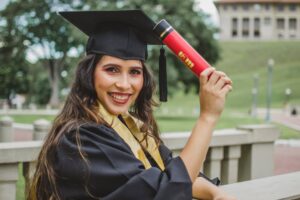 selective focus portrait photo of smiling woman in black academic dress holding diploma posing